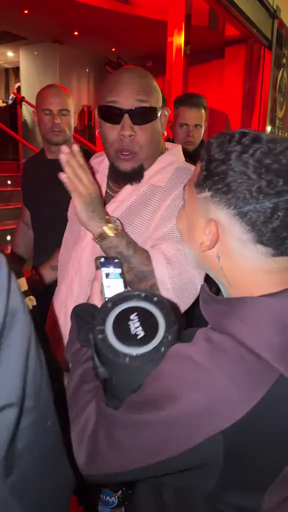 Bulin 47 in a pale-pink shirt and sunglasses facing Kevin Wai, who holds a phone and round portable speaker.